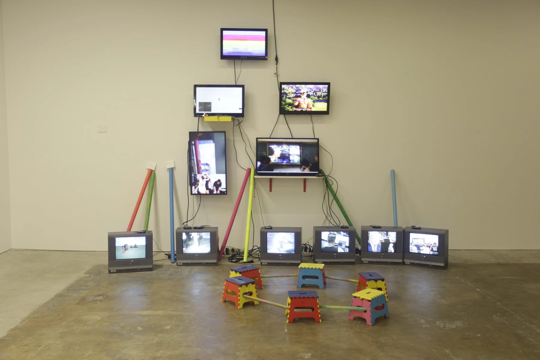 Flat screens rise up a white wall above a row of six box televisions, with black cables exposed between them. Colourful plastic stools connected by rods form a foreground circle, while red, green and blue poles lean beside the screens on a concrete floor.