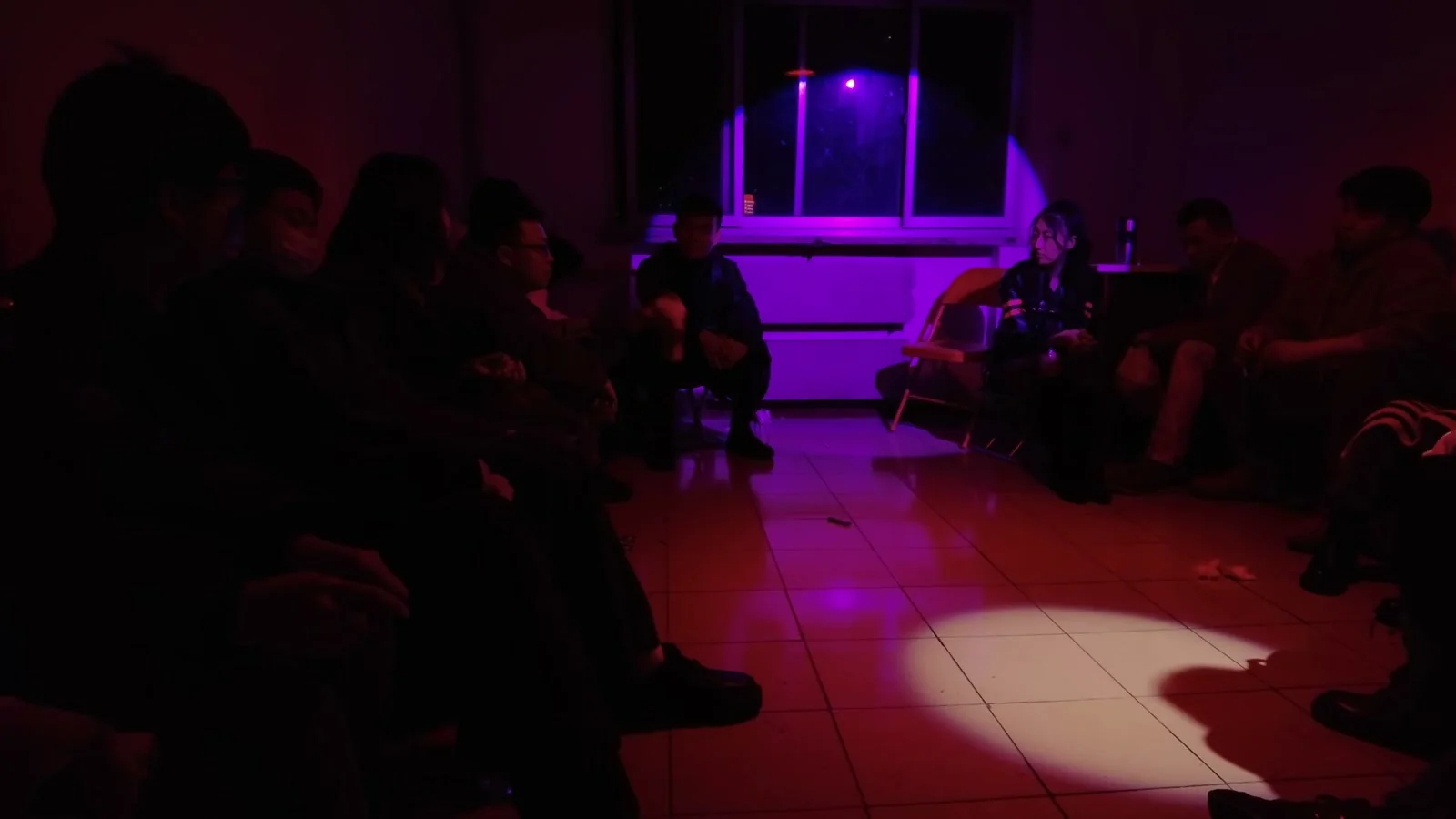 People sit along opposite sides of a dim room, leaving a tiled passage through the centre. Purple light frames the rear window and red light washes the walls, while a white circular pool of light and human shadows occupy the lower-right foreground.