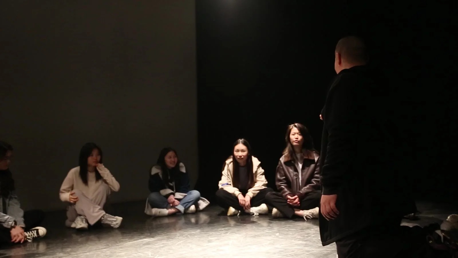 昏暗的黑色空间内，多人沿后方地面围坐，中央几张面孔被灯光照亮。右前方一名黑衣人物背对镜头站立，身体遮住部分围坐者，前景地面形成一片灰白亮区。