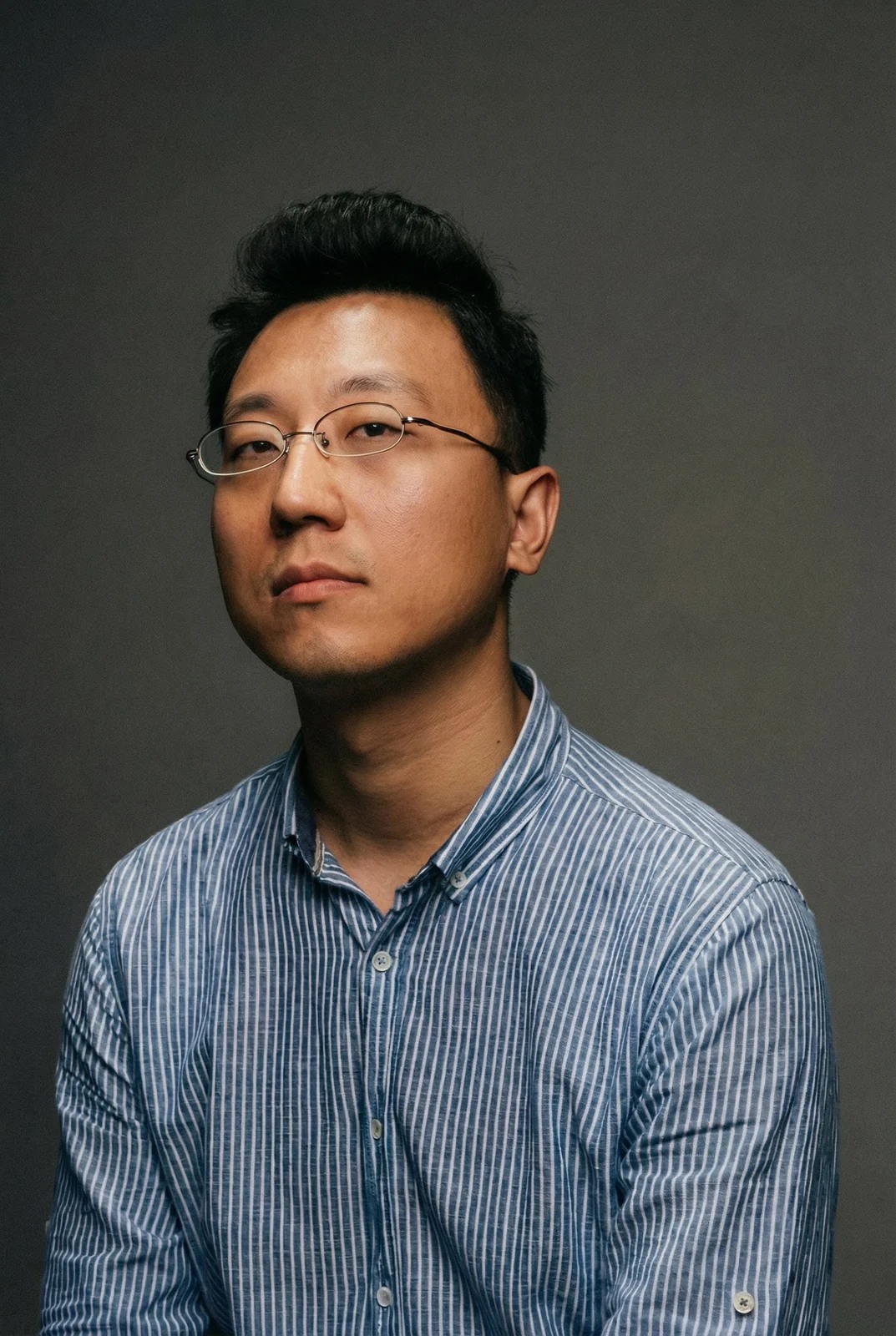灰色背景前，一名戴细框眼镜的人以半身姿态侧向坐着，脸转向镜头左侧。蓝白细条纹衬衫与深色短发清晰可见，脸部一侧较亮，背景没有其他可辨物件。