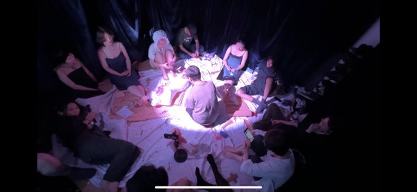 俯视画面中，人物围坐在铺有布料的中心周围，中央白紫光照亮一名人物与物件；底部保留屏幕截取的横条界面。