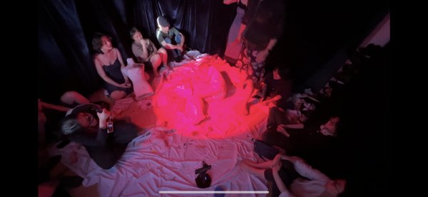 从上方拍摄的截图里，围坐者沿黑幕内侧分布，中央大块浅色布料被红光照亮。右侧黑暗中可见数张面孔，左下人物靠近画面边缘，纸张和小物件散落在布上，底部仍有白色界面横线。