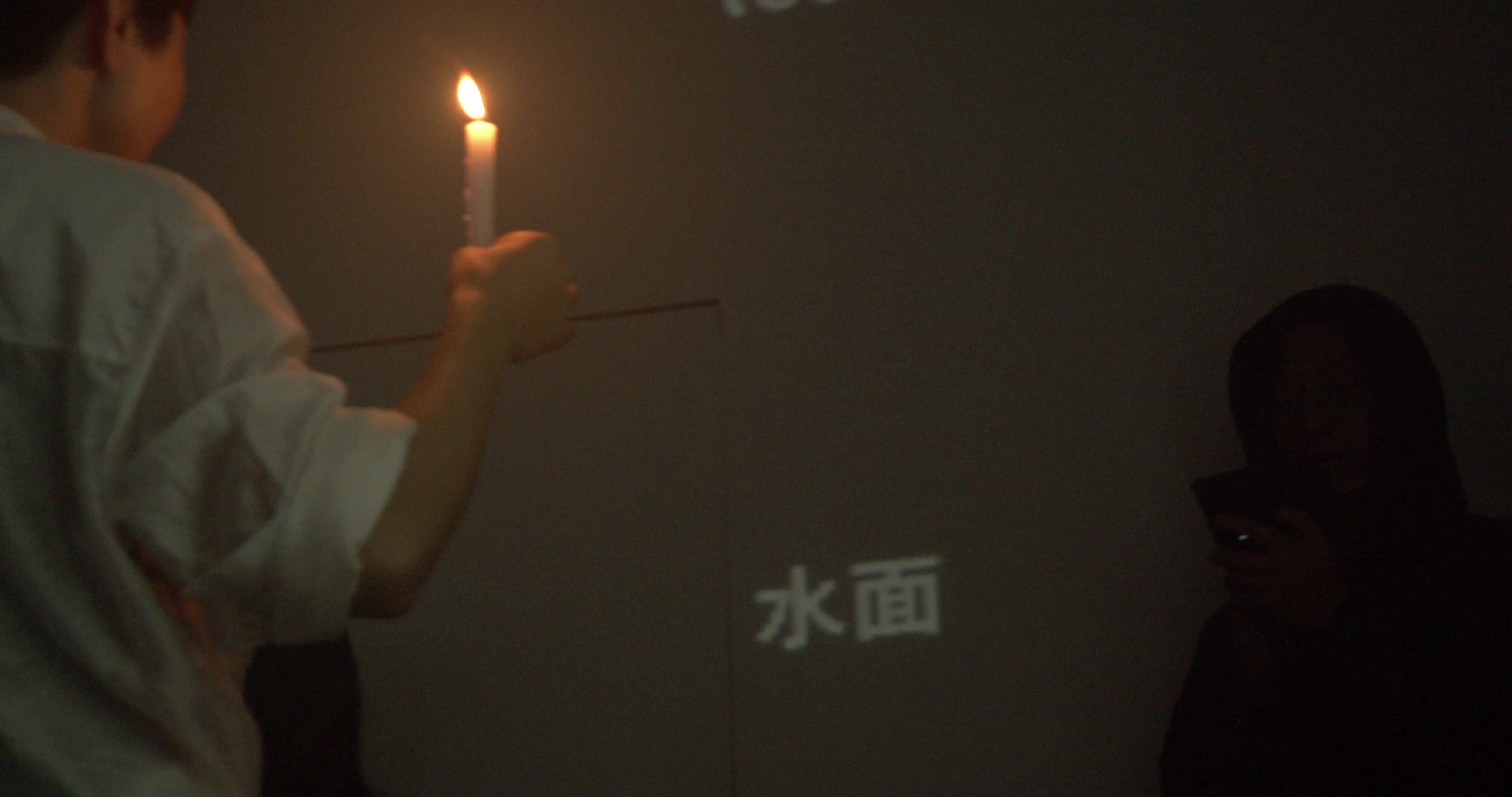 左前景穿白衣的人侧背对镜头,手中高举一支点燃的白色蜡烛,火焰成为画面最亮处。灰墙下部投出“水面”二字,右侧一人沉在暗处看着手机,近景烛光与后方文字之间留出大片空墙。