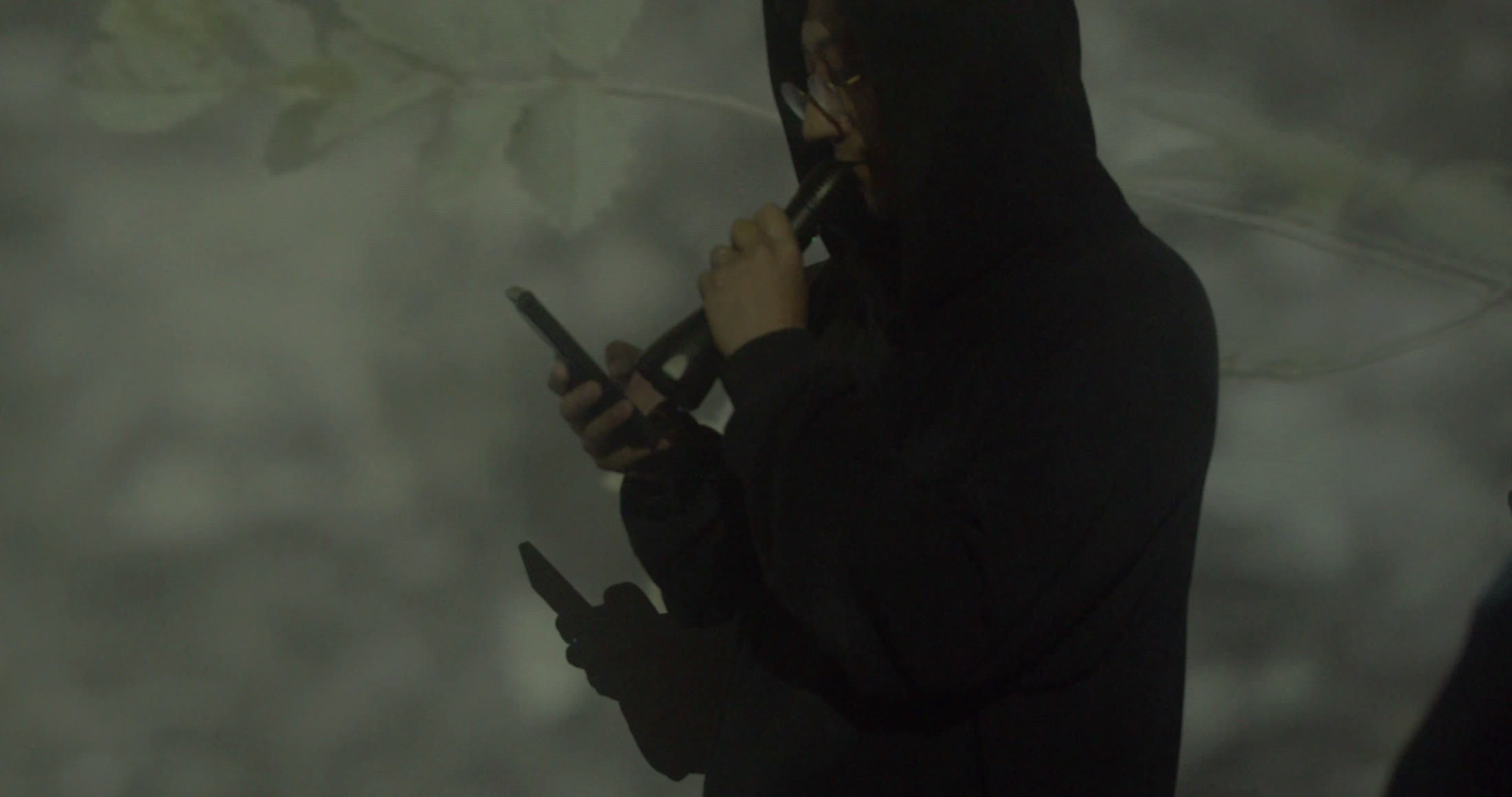 近景里，一名穿黑色连帽上衣的人低头看着手机，另一只手靠近嘴边，双臂与手机投出重叠暗影。灰绿色植物影像覆盖后墙与人物身体，右缘另有裁切的黑色轮廓，手机与手势成为主要视觉焦点。