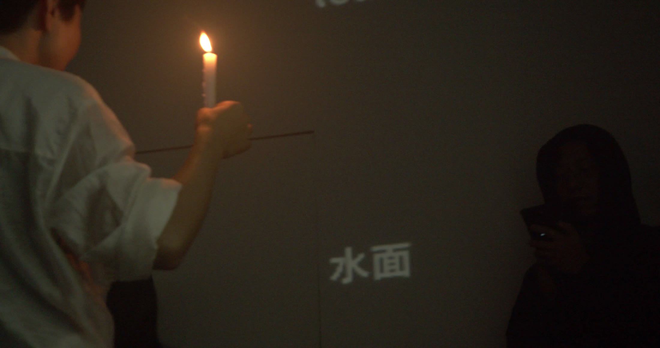 左前景穿白衣的人侧背对镜头，手中高举一支点燃的白色蜡烛，火焰成为画面最亮处。灰墙下部投出“水面”二字，右侧一人沉在暗处看着手机，近景烛光与后方文字之间留出大片空墙。