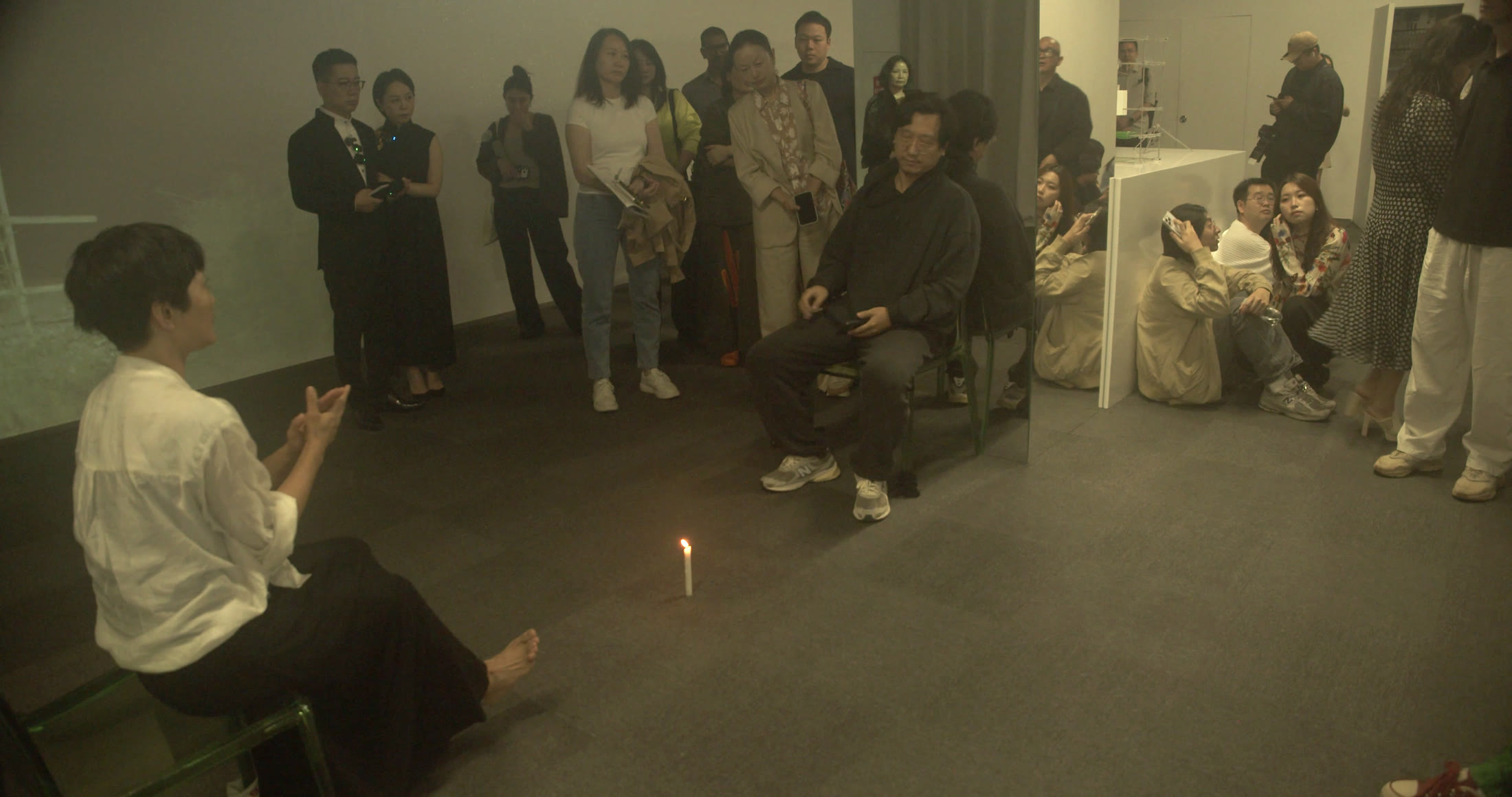 较明亮的室内全景里，左前景白衣人物坐着抬起双手，中央一名黑衣人物坐在椅上，后方多人持纸站立或在门边席地而坐。地面中央有细小烛火，投影光让白墙、灰地毯与人物之间的距离清楚展开。